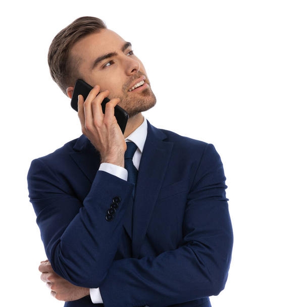 elegante jonge zakenman in marine blauw pak omhoog kijkend en pratend aan de telefoon, kruisende armen en poserend geïsoleerd op witte achtergrond in studio - Foto, afbeelding