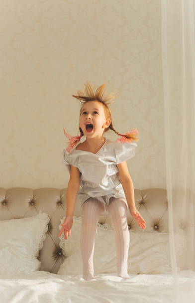 Little girl girl in a white dress with tousled hair screams and jumps on the bed. - Foto, imagen