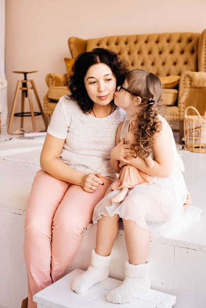 A little girl in a white dress with a soft toy in her hands is kissing her mother. Love and care concept. A loving family. Down syndrome. - Photo, Image