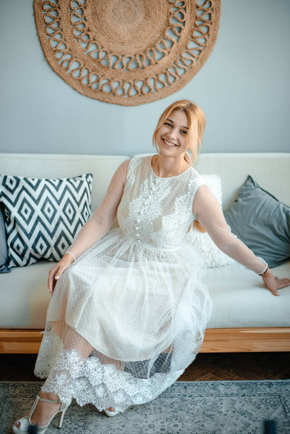 In a cozy living room, a girl in a long white dress sits on the couch while admiring. Happy bride. Wedding day. The concept of style and fashion. Copy space. Toning. - Zdjęcie, obraz