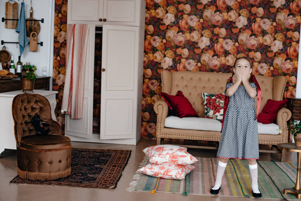A little girl in a dress and white leggings holds her face with her hands in surprise, in the background is a brown sofa with red pillows. Save space. - Φωτογραφία, εικόνα
