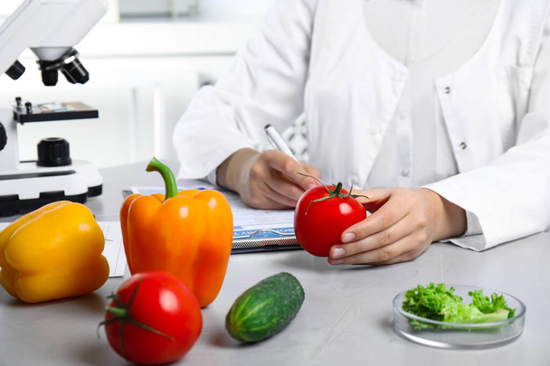 Scientist with fresh vegetables and clipboard at table in laboratory, closeup. Poison detection - Foto, afbeelding