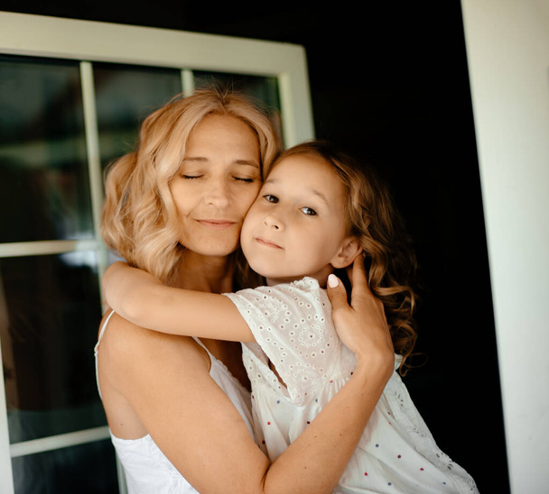 Mom blonde hugs her daughter in a white dress. The concept of happiness and love. Caring parents. Mom and daughter hugging. - Foto, immagini