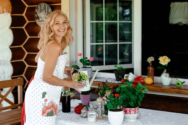A smiling girl in a white dress looks after home flowers in pots on the terrace. Flowers on the terrace. Home plant care concept. - Fotoğraf, Görsel