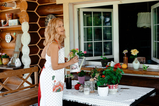A blonde girl in a white dress and an apron holds a watering can in her hands, watering houseplants. Home plant care concept. flowers on the terrace. - Photo, Image