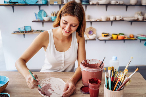 Eine attraktive junge Künstlerin schafft in ihrem eigenen Atelier. Handgefertigte Produkte zum Verkauf. Dekoriert den Teller mit aufgeklebten Kaffeebohnen. Geräumiger, heller Raum mit Fertigprodukten im Hintergrund. - Foto, Bild