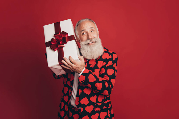 Photo of funny bearded old man hold giftbox prepare unpack make guess wear suit isolated red background - 写真・画像