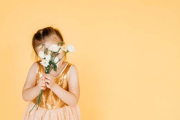A little girl in a dress stands in a room on a yellow foil holding white flowers in her hands. Helping children with special needs. Copy space. - Фото, зображення