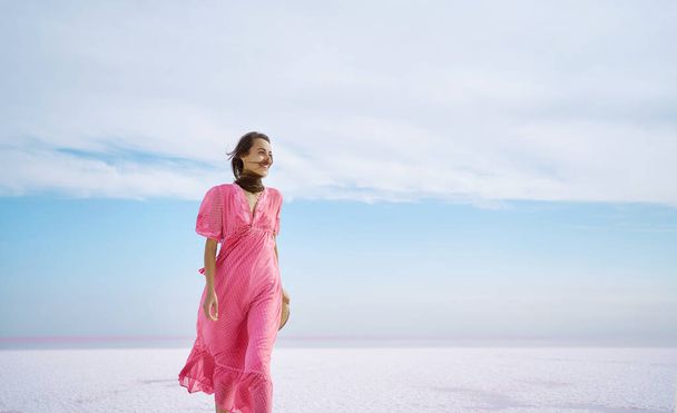 Freedom cheerfulness woman walking on white salty beach on pink salt lake with paradise view and beautiful blue sky. Elegant girl in blowing by wind pink dress - Photo, image