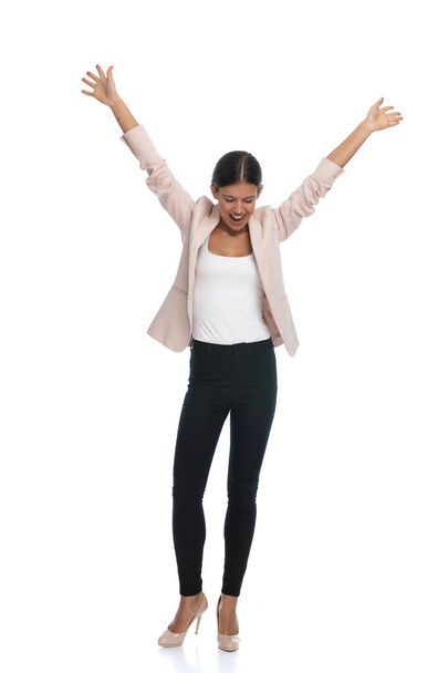 full body picture of happy businesswoman in pink jacket holding arms in the air, looking down and laughing, standing and posing isolated on white background in studio - Photo, Image
