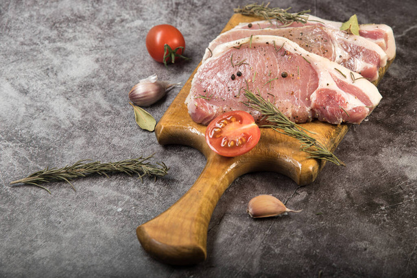 Carne fresca, carbonato de cerdo en una tabla de cortar con tomates y especias sobre un fondo gris moteado - Foto, Imagen