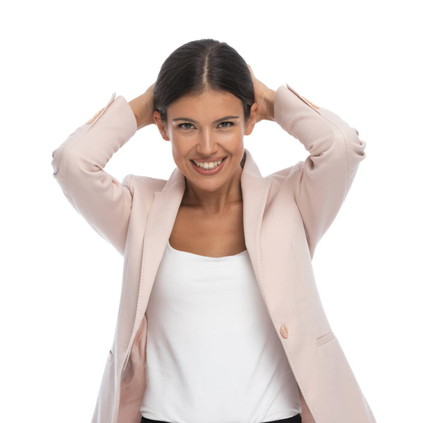 χαρούμενη έξυπνη περιστασιακή γυναίκα που κρατάει τα χέρια πίσω από το κεφάλι και χαμογελά, φοράει ροζ μπουφάν και στέκεται απομονωμένη σε λευκό φόντο στο στούντιο - Φωτογραφία, εικόνα