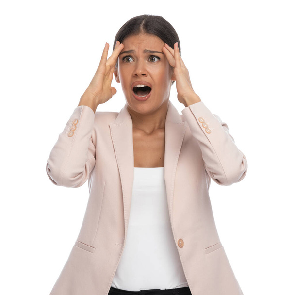 mujer joven perturbada en chaqueta rosa cogida de la mano a la frente, gritando, teniendo un dolor de cabeza y mirando a un lado sobre fondo blanco en el estudio - Foto, imagen
