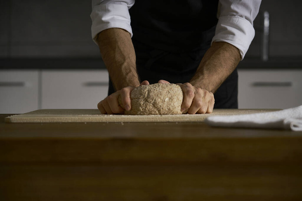 The hands of the strong man knead the dough from which they will make bread, pasta, cake or pizza. In his kitchen he carries on the tradition of homemade pasta. A cloud of white flour flies like dust. - Foto, Imagen
