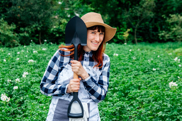 Portret pięknej farmerki z łopatą uśmiechniętą do aparatu w słomkowym kapeluszu, otoczonej świeżymi zielonymi liśćmi wielu roślin w ogrodzie warzywnym, z zielenią w tle. - Zdjęcie, obraz