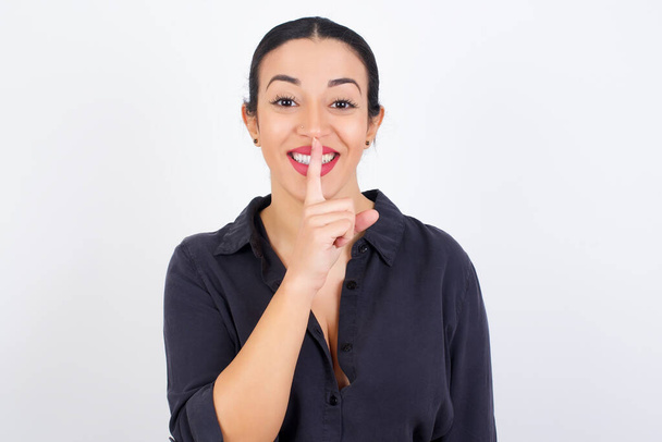 Mujer árabe sonriente usando vestido contra fondo de estudio blanco hace gesto de silencio, sostiene el dedo por encima de los labios se esconde secreto. Silencio, por favor.. - Foto, imagen