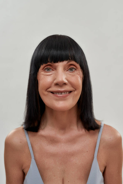 Feeling so happy. Close up portrait of beautiful cheerful senior woman smiling at camera isolated over grey background, posing half naked in studio - Foto, Imagem