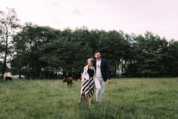 Pohledný chlap a blondýnka kráčející po trávě u řeky a lesa. Drží se za ruce a usmívají se na pozadí pastvin s krávami a koňmi - Fotografie, Obrázek