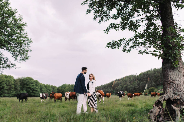 Handsome guy and blonde girl walking on the grass near the river and forest. They hold hands and smile on the background of pasture with cows and horses - Photo, Image