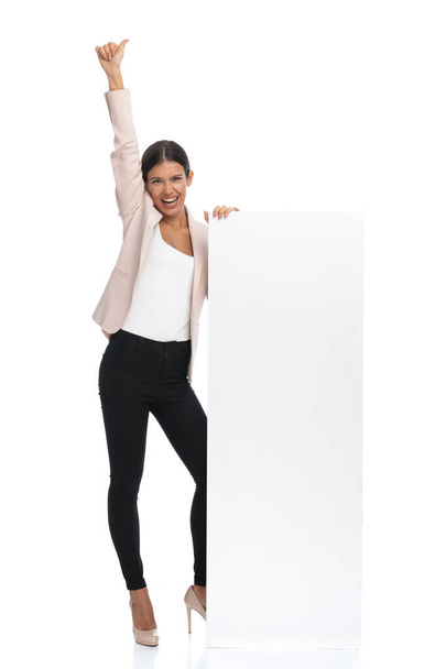 full body picture of happy woman in pink jacket standing next to empty board, holding arms in the air and making thumbs up gesture on white background in studio - Photo, Image