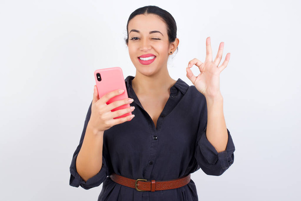 Mujer árabe hermosa feliz vistiendo vestido contra fondo de estudio blanco enviando un mensaje en su teléfono inteligente o tomando una selfie y haciendo señal de ok con su mano. - Foto, imagen