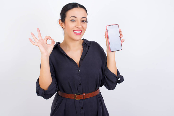 Ενθουσιασμένη Άραβας γυναίκα φορώντας φόρεμα κατά λευκό φόντο στούντιο δείχνει το smartphone κενή οθόνη, αναβοσβήνει το μάτι και κάνει ok υπογράψει με το χέρι. Έννοια διαφήμισης. - Φωτογραφία, εικόνα