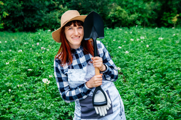 Elinde kürekle kameraya gülümseyen güzel bir kadın çiftçi portresi. Hasır şapka takıyor ve arka planda yeşillik ve sebze bahçesindeki birçok bitkinin taze yeşil yapraklarıyla çevrili.. - Fotoğraf, Görsel