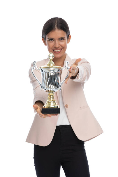 гордая деловая женщина в розовой куртке, показывая серебряный трофей, представляя и хвастаясь, улыбаясь и позируя изолированы на белом фоне в студии - Фото, изображение
