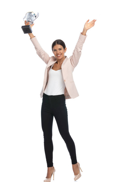 full body picture of smiling young woman in pink jacket holding hands up, holding trophy and smiling on white background in studio - Photo, Image