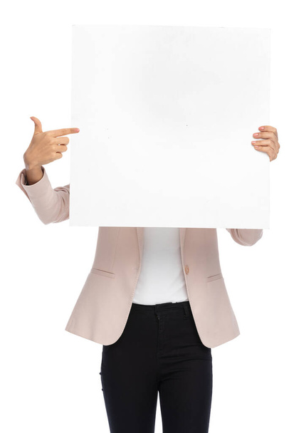 portrét atraktivní mladé ženy v růžové bundě zakrývající tvář s prázdnou deskou a ukazujícími prsty, ukazující a prezentující na bílém pozadí ve studiu - Fotografie, Obrázek