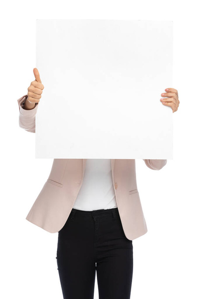 smart casual woman in pink jacket covering face with empty board and making thumbs up gesture on white background in studio - Photo, Image