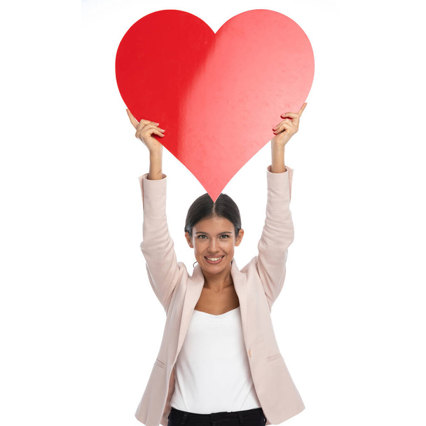 proud young female model in pink jacket holding big red heart above head and celebrating valentine's day, standing and posing isolated on white background in studio - Photo, Image
