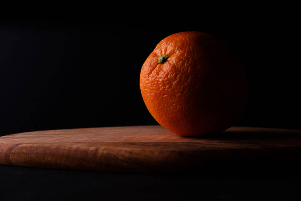 Częściowo oświetlona pomarańcza na drewnianej desce z czarnym tłem. Koncepcja ciemnego jedzenia. Koncepcja wegańska. - Zdjęcie, obraz