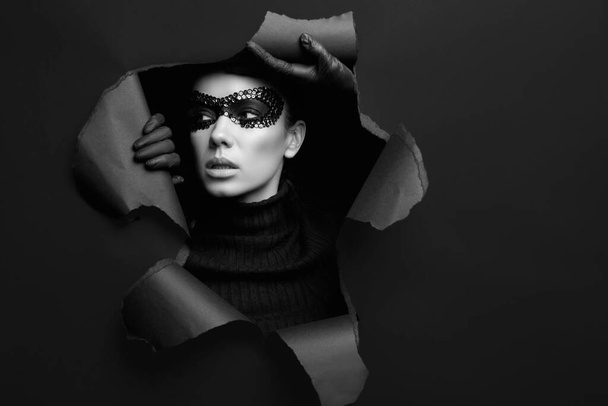 Portrait of charming elegant brunette woman in black turtleneck sweater and sequins mask looks through a ripped studio background - Fotoğraf, Görsel