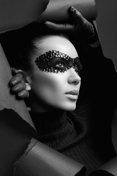 Portrait of charming elegant brunette woman in black turtleneck sweater and sequins mask looks through a ripped studio background - Photo, Image