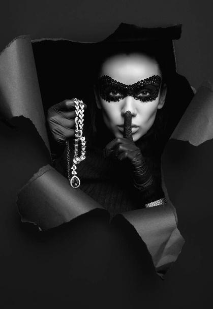 Retrato de encantadora mujer morena elegante en jersey de cuello alto y máscara de lentejuelas con guantes negros posando con un collar de diamantes robado - Foto, imagen