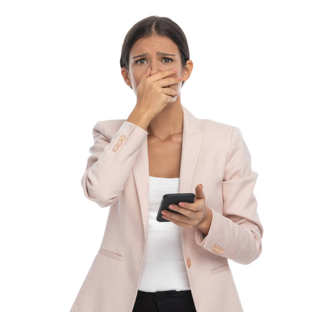 sad young businesswoman in pink jacket making an unhappy face, covering mouth with hand and reading texts on white background in studio - Foto, Imagem