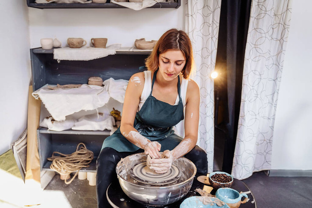 Профессиональными движениями рук, женщина-гончар готовит кусок глины на гончарном круге, разминает глину и выкатывает ее, чтобы сделать миску, вазу, кувшин, тарелку. Гончарная мастерская. Молодой специалист. - Фото, изображение