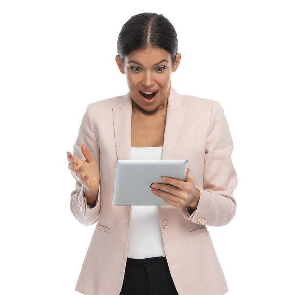 portrait of shocked young female in pink jacket on white background, holding tab and reading emails, making a surprised face in studio - Valokuva, kuva