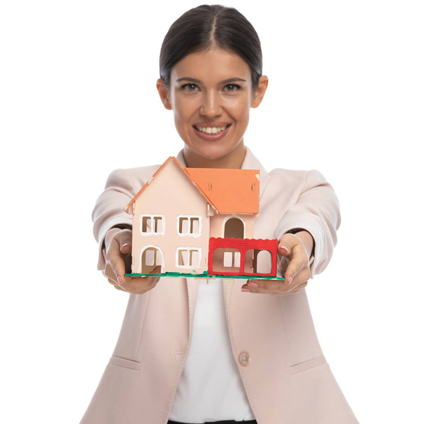 усміхнена розумна випадкова жінка в рожевій куртці тримає модель будинку і рекомендує, стоячи і позує на білому тлі в студії
 - Фото, зображення