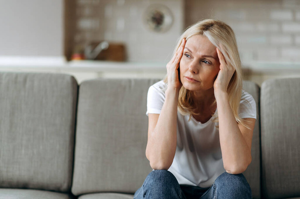 Surullinen valkoihoinen vanhempi nainen päällään rento vaatteita istuu sohvalla kotona yksin tuntuu onneton, koska päänsärky, stressi, sairaus tai huonoja uutisia, hän tarvitsee lepoa ja unta - Valokuva, kuva
