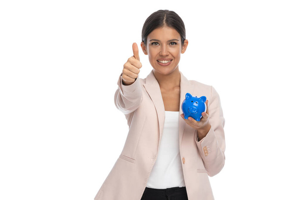 attractive brunette woman in pink jacket making thumbs up gesture, smiling and showing piggy bank on white background in studio - Photo, Image