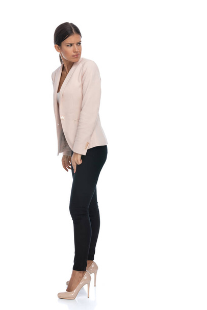 πλαϊνή άποψη της ελκυστική επιχειρηματίας σε ροζ σακάκι ποζάρουν σε λευκό φόντο, κοιτάζοντας πάνω από τον ώμο και συνοφρύωμα, στέκεται στο στούντιο - Φωτογραφία, εικόνα