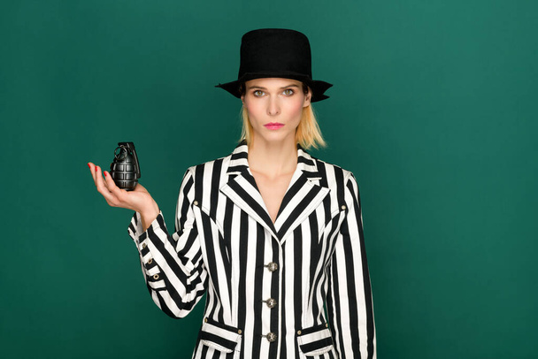 Modèle blond élégant et confiant en veste rayée noire et blanche et chapeau tenant une grenade noire sur fond vert - Photo, image