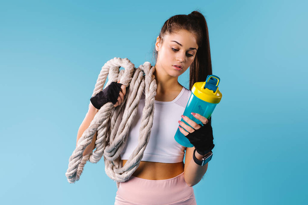 Sporty girl with a rope on her shoulder and with a protein shake in her hands. Blue background and side space. - Фото, изображение