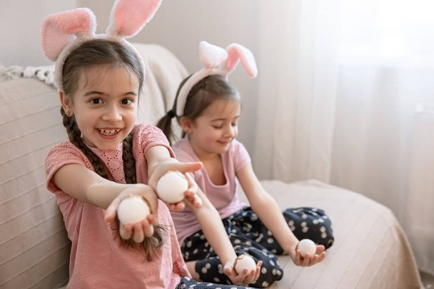 Маленькі сестри з вухами кролика і великодніми яйцями позують для камери на дивані вдома. Концепція Великодня фотосесії
. - Фото, зображення