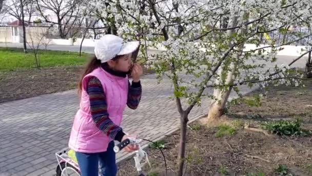 Une fille avec un vélo et une veste rose sans manches renifle des fleurs sur une branche dans un parc printanier ensoleillé. - Séquence, vidéo