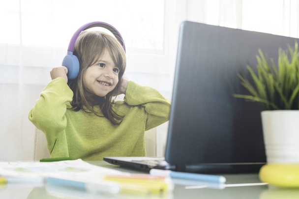 Niña de la escuela primaria en auriculares sentados en el escritorio en el ordenador portátil - Sonriendo chica linda estudiando en línea desde casa y aprendiendo usando lecciones de Internet durante el brote de coronavirus - Concepto de educación en el hogar - Foto, imagen