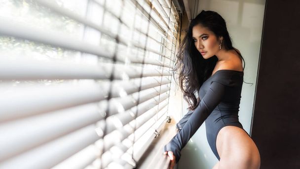 Sexy brunette Aziatische vrouw poseren in de buurt venster met natuurlijk licht door gordijn. Sensueel meisje in topvorm. Mode schoonheid en kijk met kopieerruimte voor tekst. - Foto, afbeelding