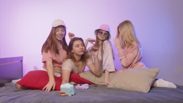 Cuatro hermosas mujeres jóvenes felices en pijama se sienta en la cama y toma un silfie en la cámara vintage en gafas de sol y sombreros en la despedida de soltera - Metraje, vídeo